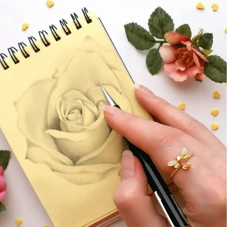 Hand holding a pen over a notebook with a rose drawing, surrounded by flowers and decorative elements.