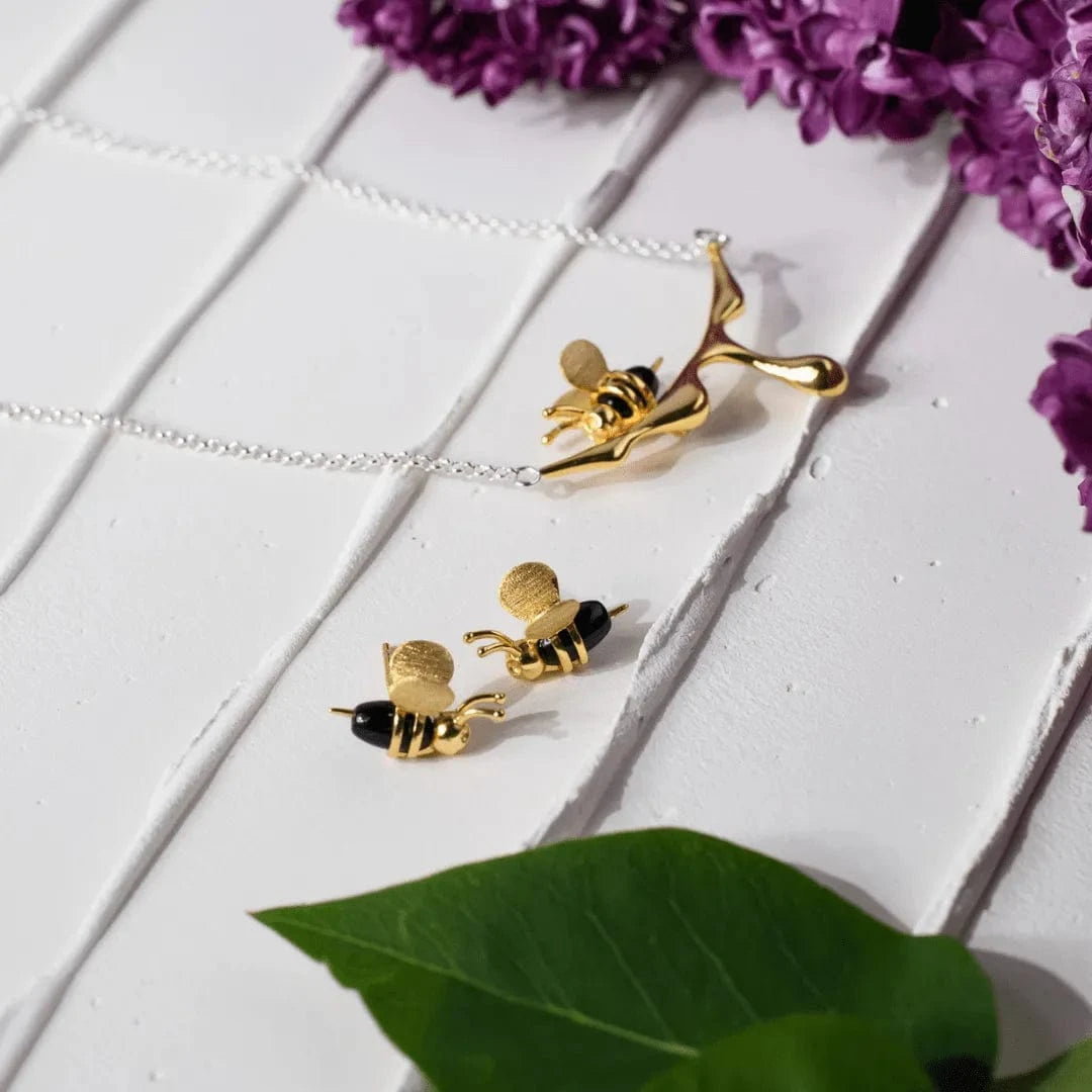 Gold bee-shaped earrings on a white surface with purple flowers in the background