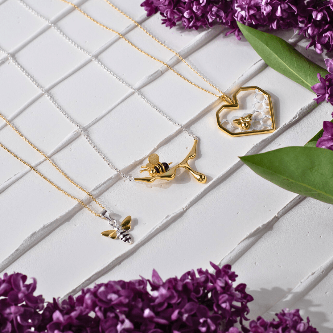 Gold bee necklaces on a white textured surface with purple flowers.