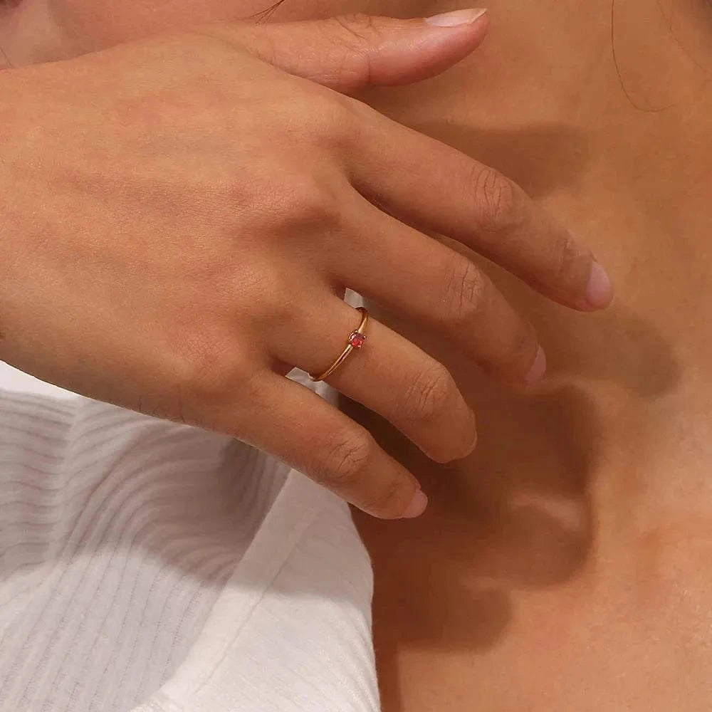 Close-up of a hand wearing a gold ring with a red stone on a neutral background