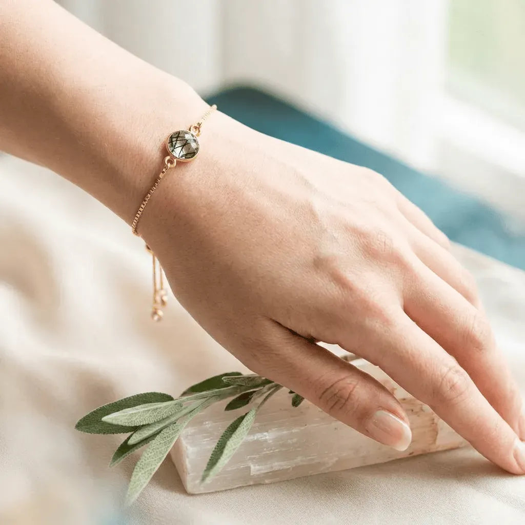 Hand wearing a gold bracelet with a green stone, resting on a crystal and leaf.
