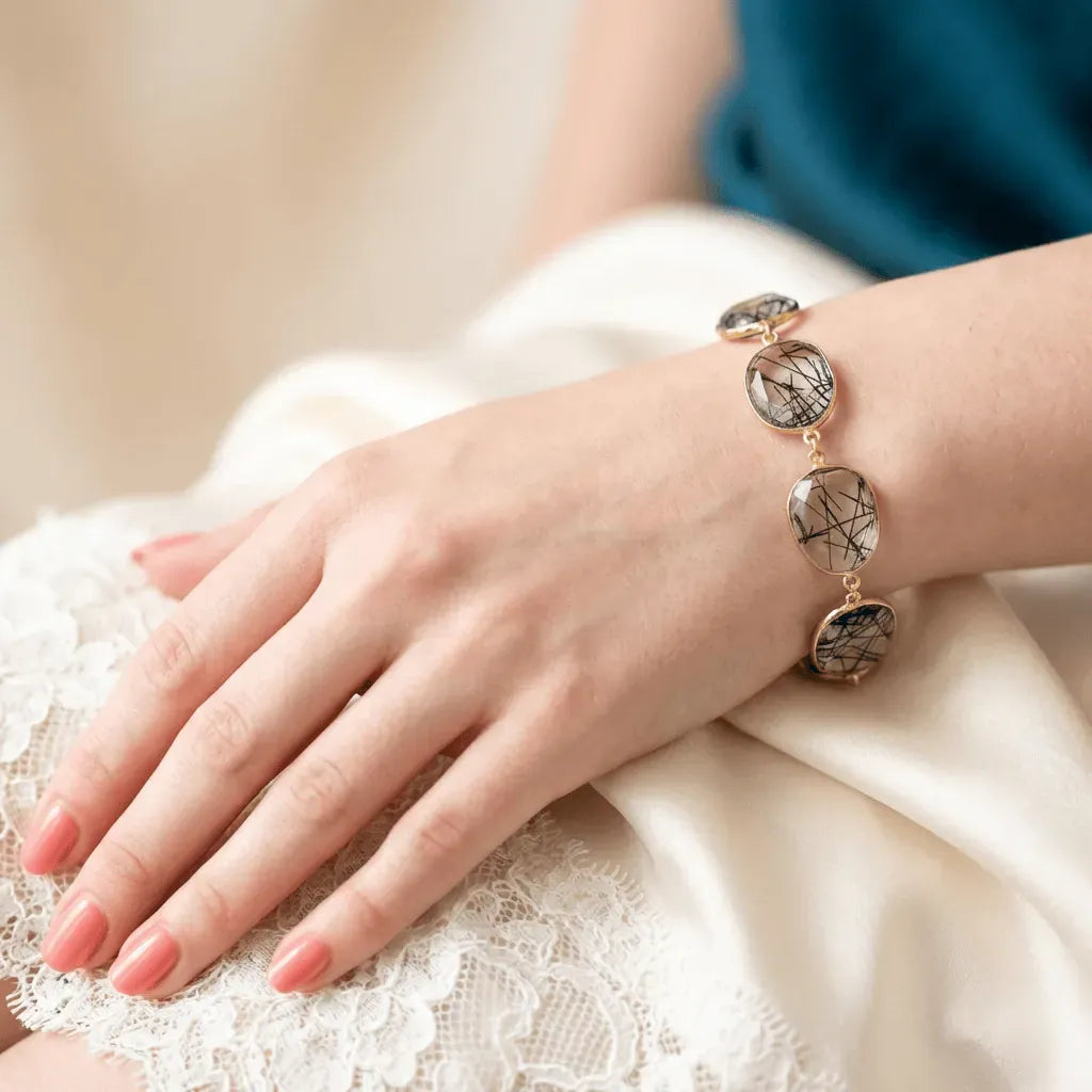 Hand wearing a bracelet with gold accents and gemstones on a white lace fabric background
