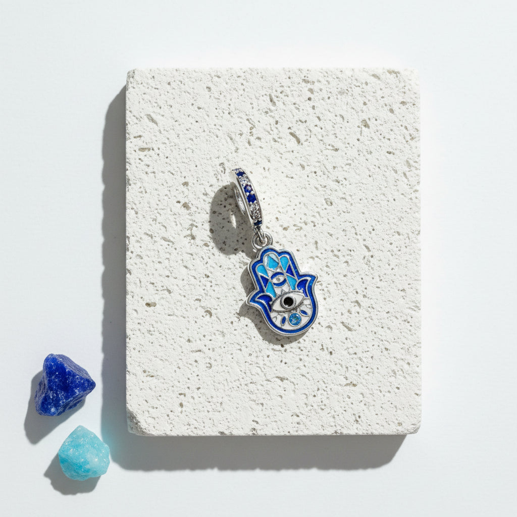 Blue enamel hamsa hand pendant with eye detail on a sterling silver clip bail, on white stone beside blue crystals