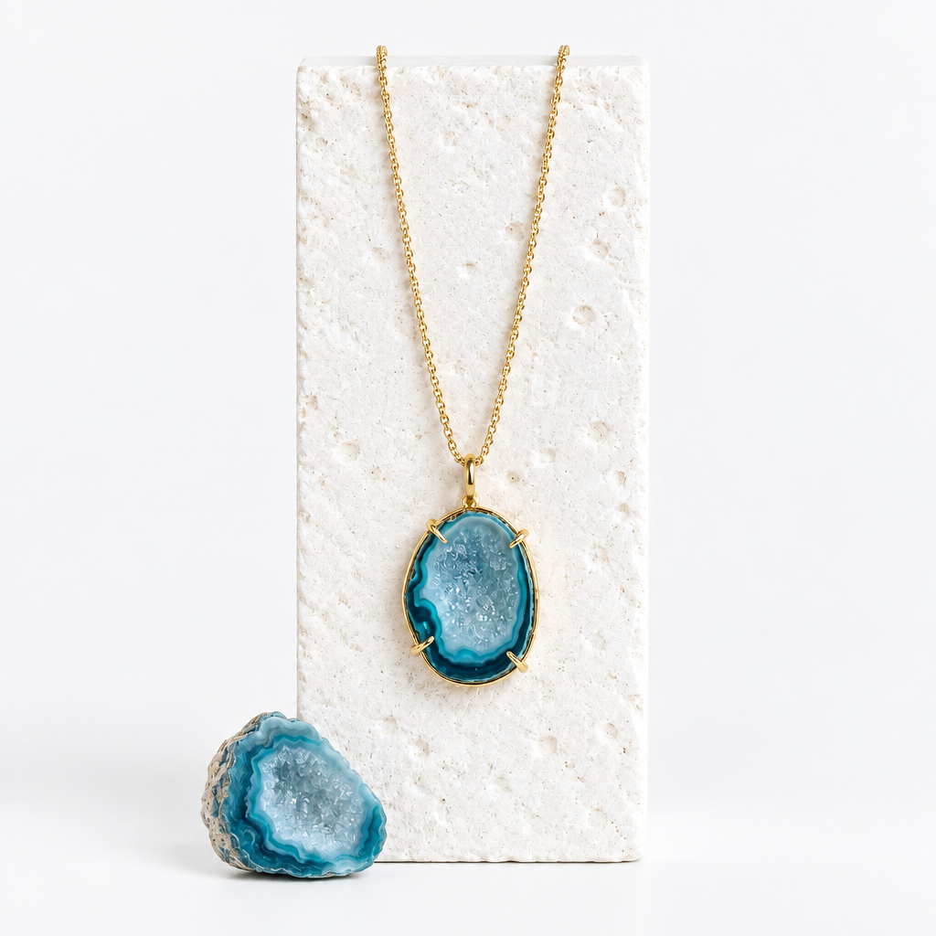 Blue druzy agate pendant necklace hanging against a textured white card with a raw blue crystal below