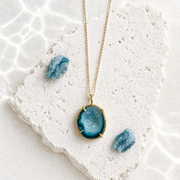 Blue druzy agate pendant necklace, a free-form stone in a gold-plated bezel, lying on white stone beside raw blue crystals