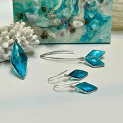 Blue square earrings with silver hooks on a white surface, with a decorative blue and white background.