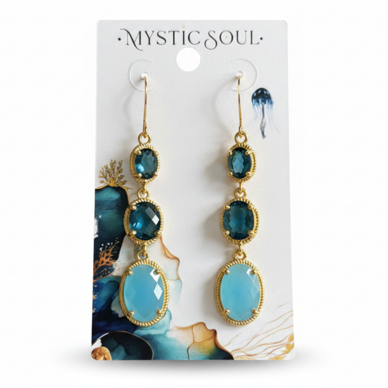 A pair of Mystic Soul drop earrings with blue faceted gemstones in gold settings, displayed on a white branded card against a neutral gray background.