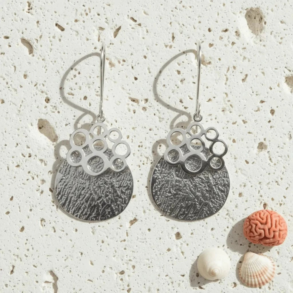 Pair of silver earrings on a textured surface with small shells and a coral object.