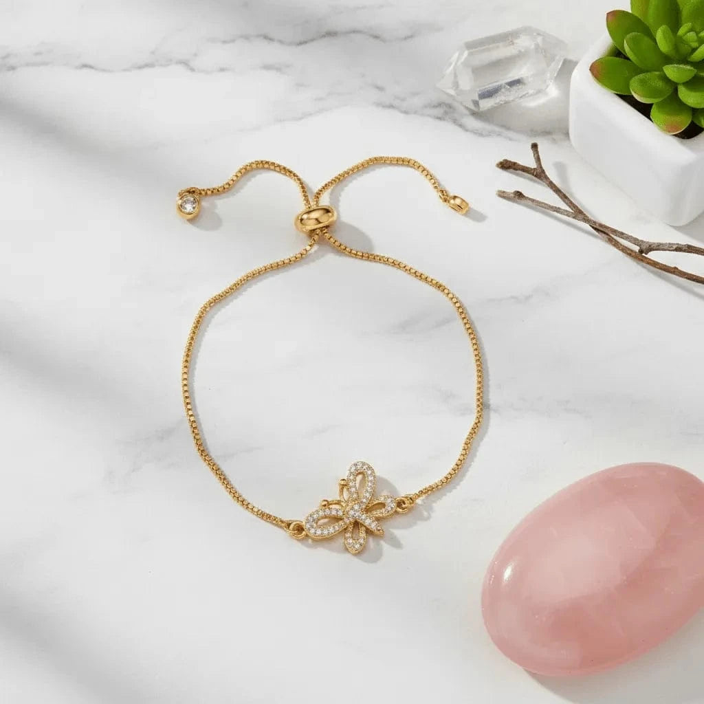 gold bracelet with butterfly charm on a white surface with a pink stone and plant in the background
