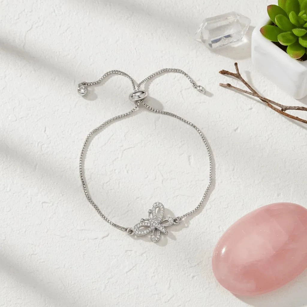 Silver bracelet with butterfly charm on a white surface with a pink stone and plant in the background