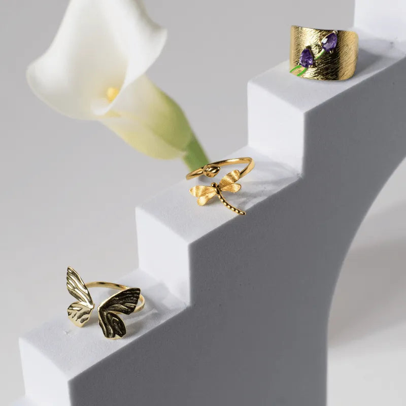 Gold rings with floral and butterfly designs on a white surface with a white flower.