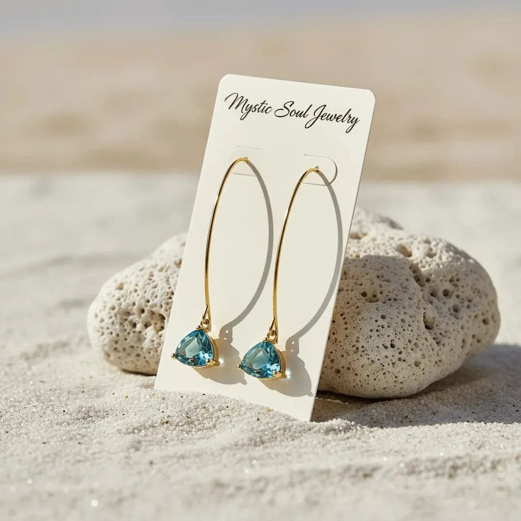 Gold earrings with blue gemstones on a 'Mystic Soul Jewelry' card, placed on sand with rocks.