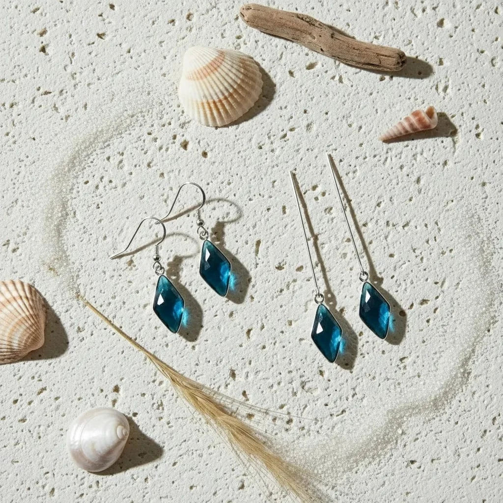 Blue teardrop earrings on a sandy surface with shells and driftwood.