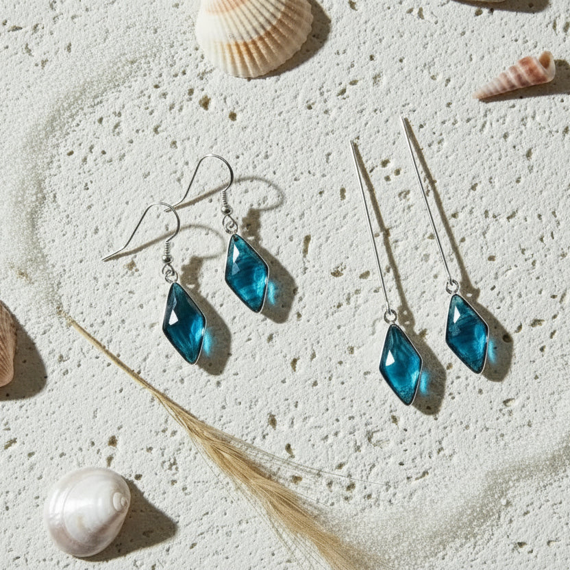 Blue teardrop earrings on a sandy surface with shells and driftwood.