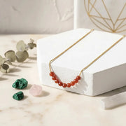 carnelian_necklace_gold beads on a white stand with green and pink stones on a light surface
