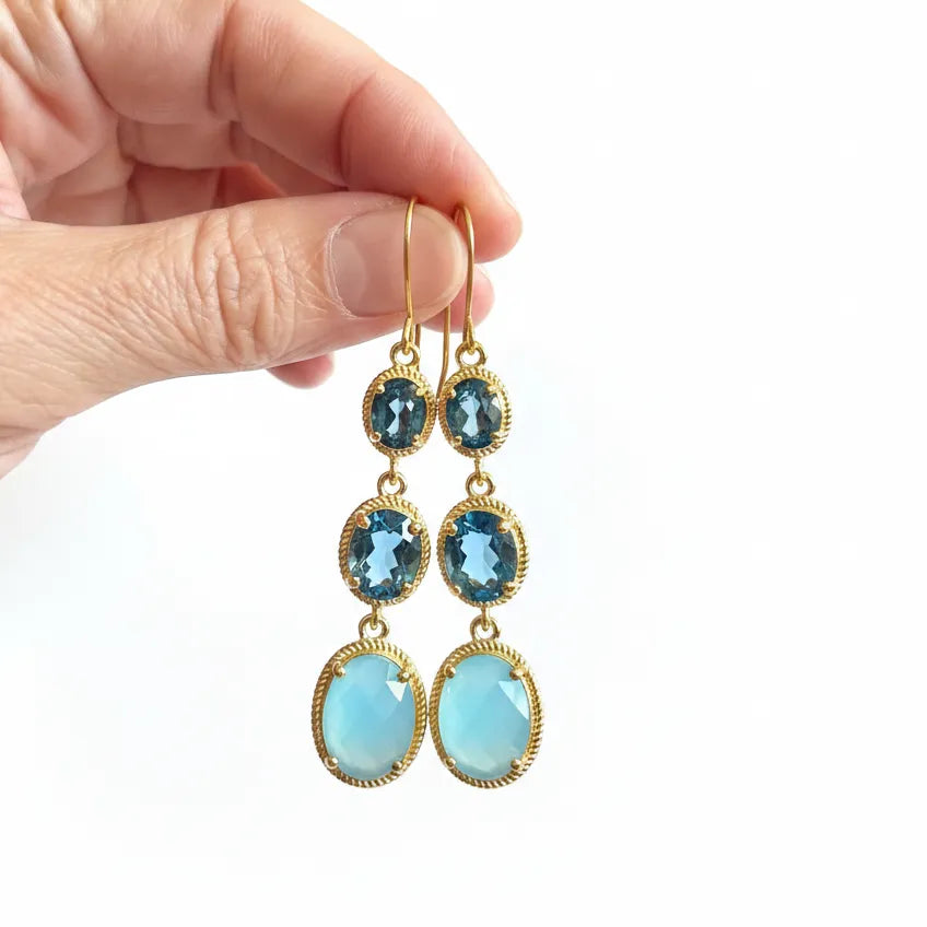 A pair of gold drop earrings with blue gemstones, held by a hand against a plain white background.