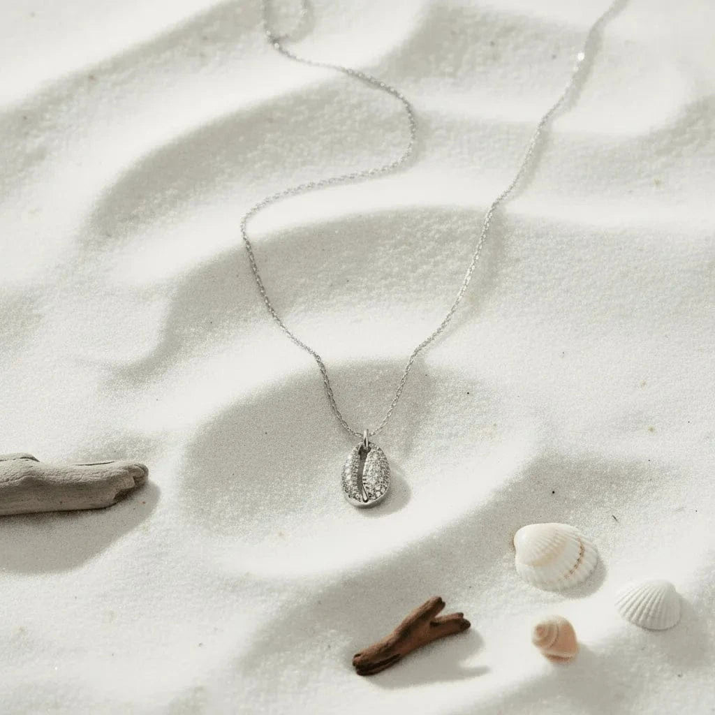 Silver necklace with a pendant on a sandy surface with driftwood and shells.
