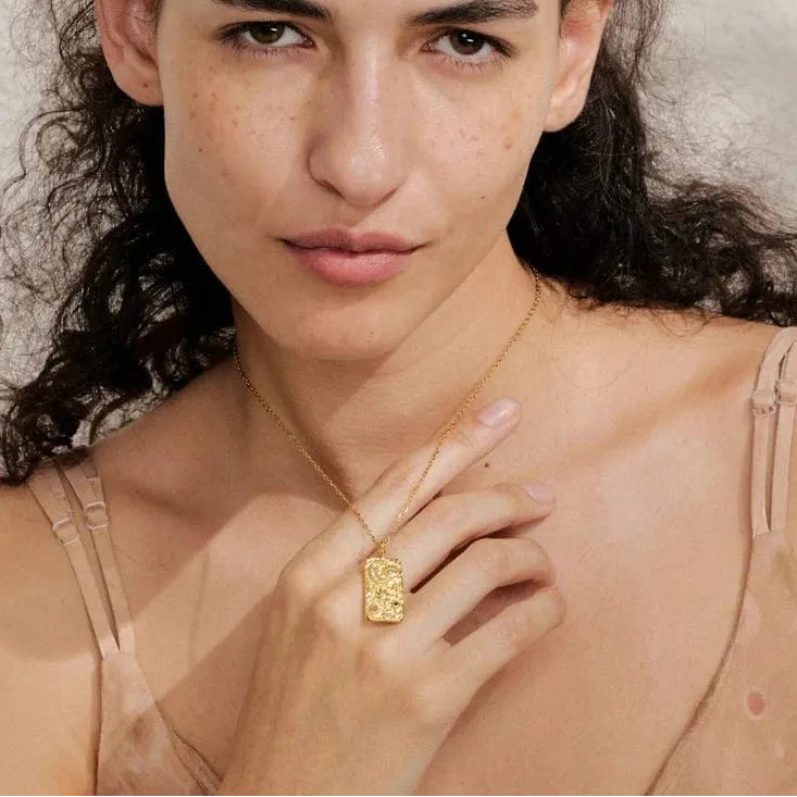 Woman wearing a gold necklace with a textured pendant against a neutral background