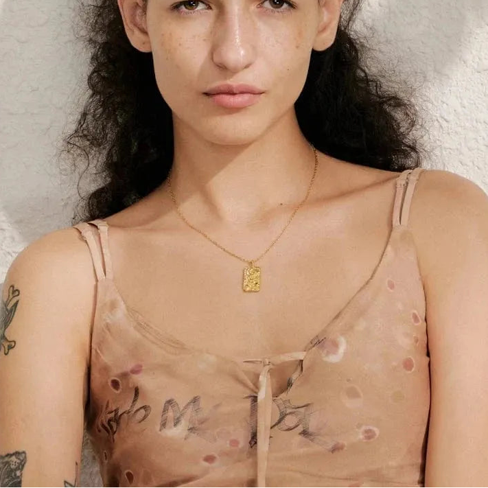 Woman wearing a sheer top with floral patterns and a gold necklace against a neutral background