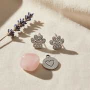 Silver paw-shaped earrings and a heart pendant on a beige surface with a pink stone and lavender flower.