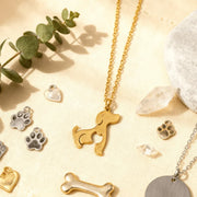 Gold and silver pet-themed necklaces and pendants on a beige surface with greenery.
