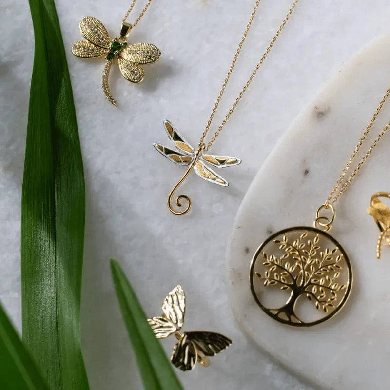 Gold jewelry with dragonfly, butterfly, and tree of life designs on a marble surface.