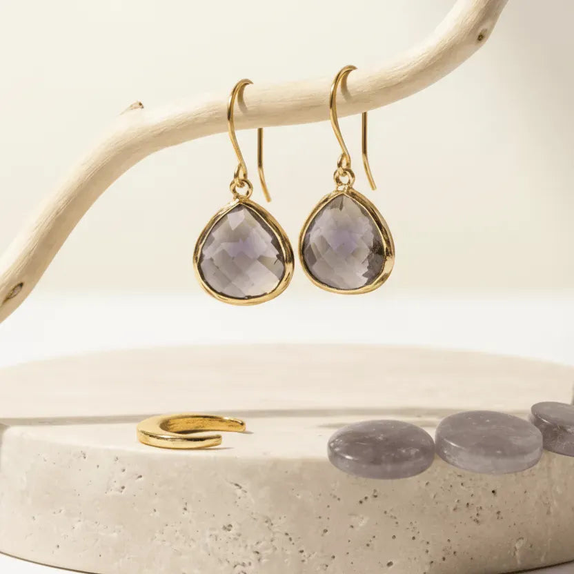 Gold earrings with teardrop-shaped gemstones on a beige stand with a branch background