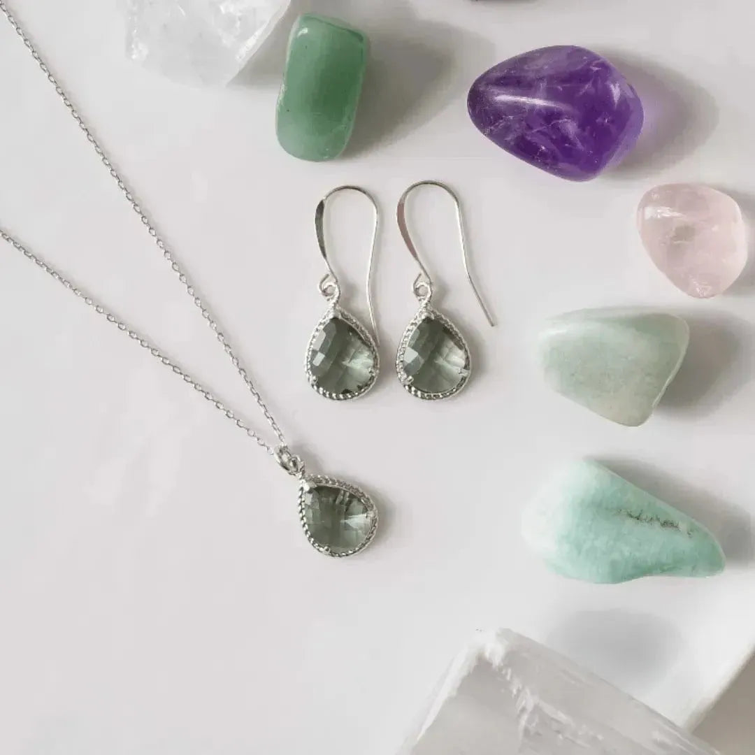 Silver jewelry set with green gemstone pendant and earrings, surrounded by colorful crystals on a light background.