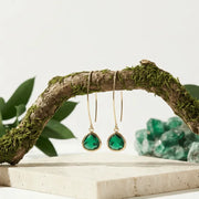 Green teardrop earrings hanging from a branch with green stones in the background