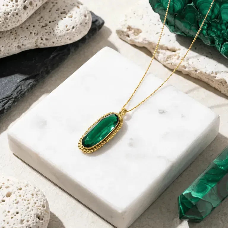 Gold necklace with a green pendant on a white surface, surrounded by green crystals and stones.