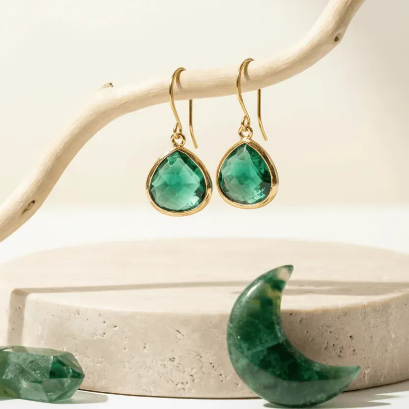 Gold earrings with green gemstones on a natural wood branch and stone surface.