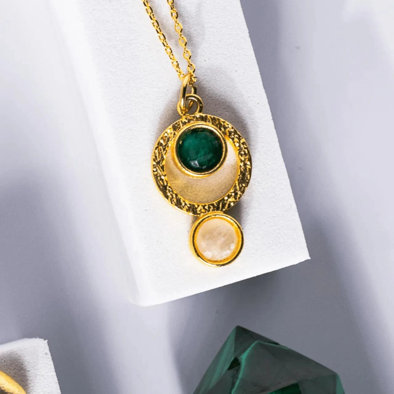 Gold necklace with green and beige pendant on a white card against a gray background