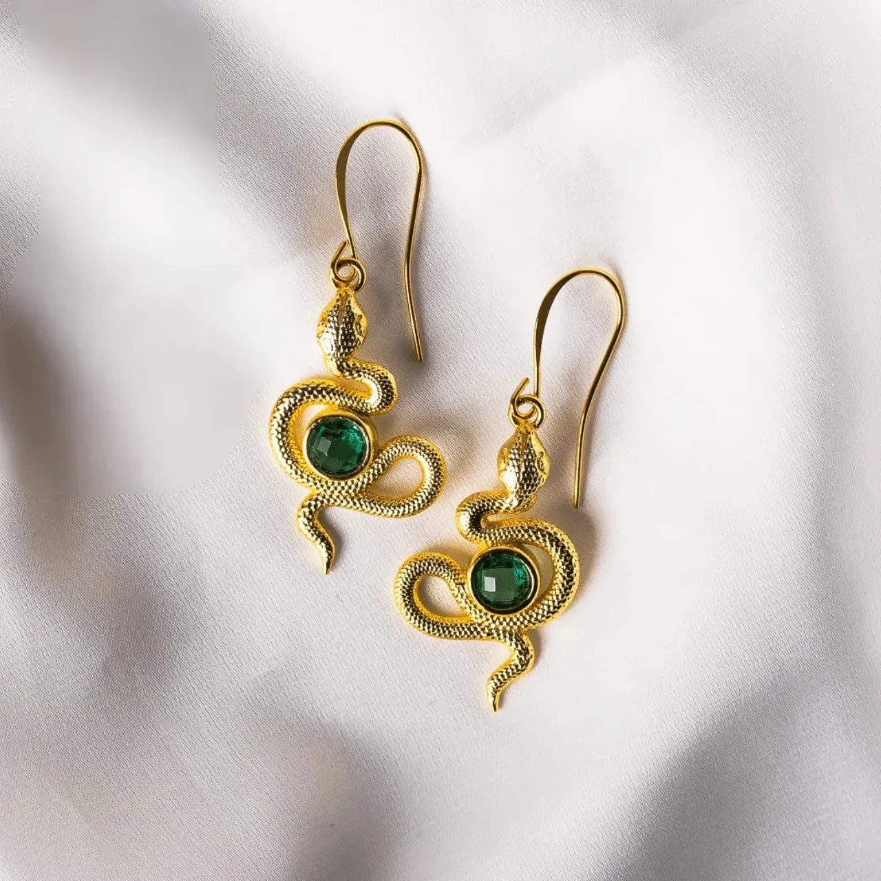 Gold snake-shaped earrings with green gemstones on a white fabric background
