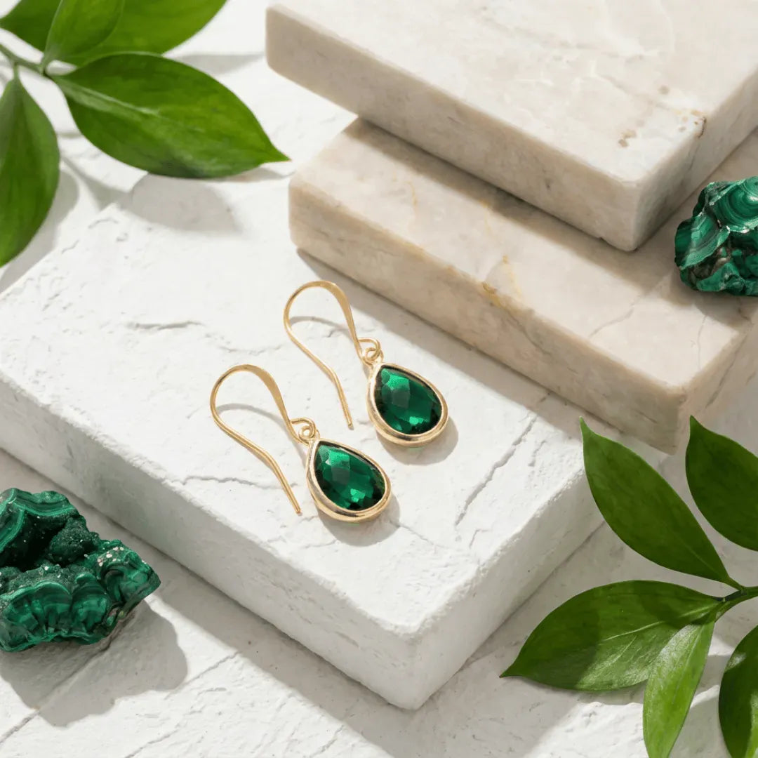 Gold earrings with green gemstones on a white surface with green leaves.