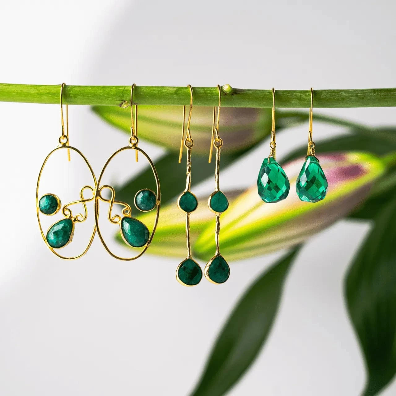 Gold earrings with green teardrop beads hanging on a branch with leaves in the background