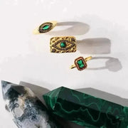 Three gold rings with green gemstones on a white surface with green fabric underneath.