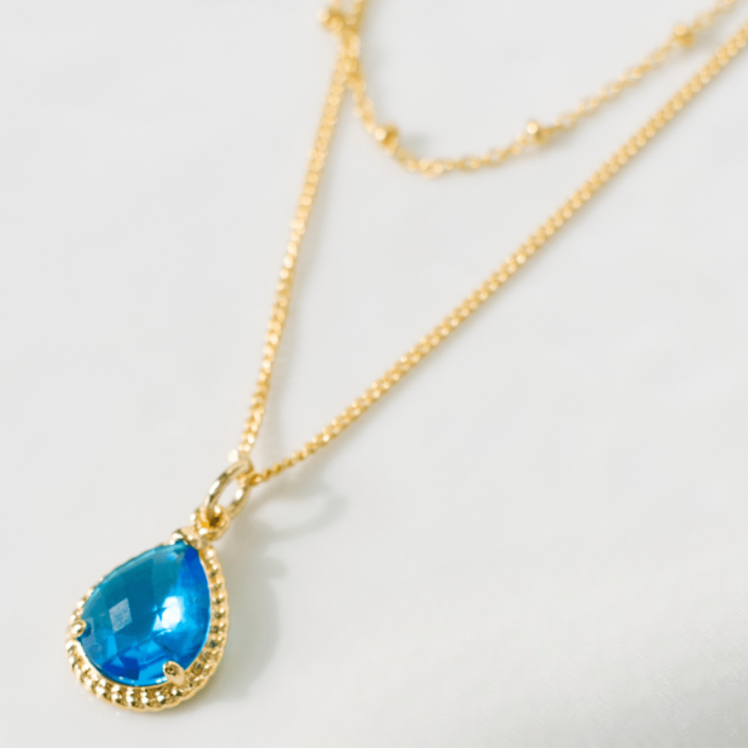 Gold necklace with a blue teardrop pendant on a light gray background