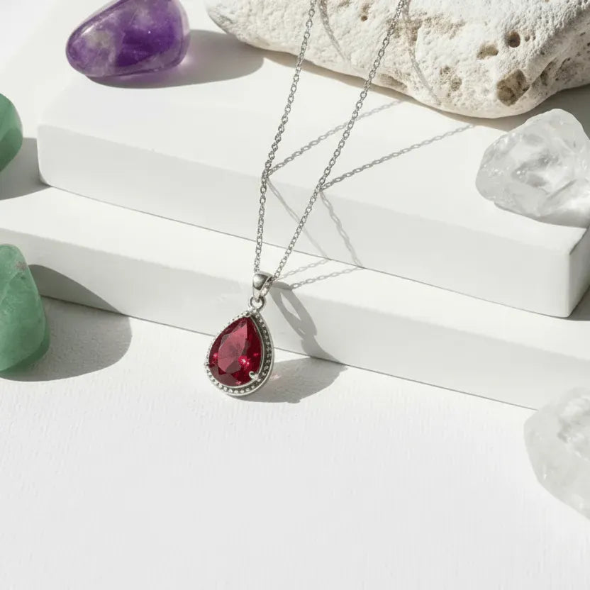 Necklace with a red teardrop pendant on a white surface with crystals and stones.