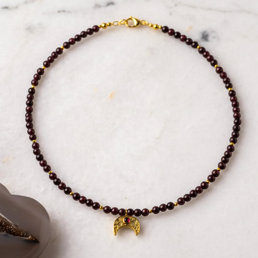 A beaded necklace with a half-moon pendant, displayed on a white surface, alongside a black and a gray stone with a crystal in the center.