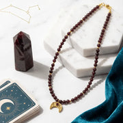 Garnet Moon Necklace on a white surface with cards and a crystal, featuring text about the necklace's symbolism and connection to the moon's cycles.