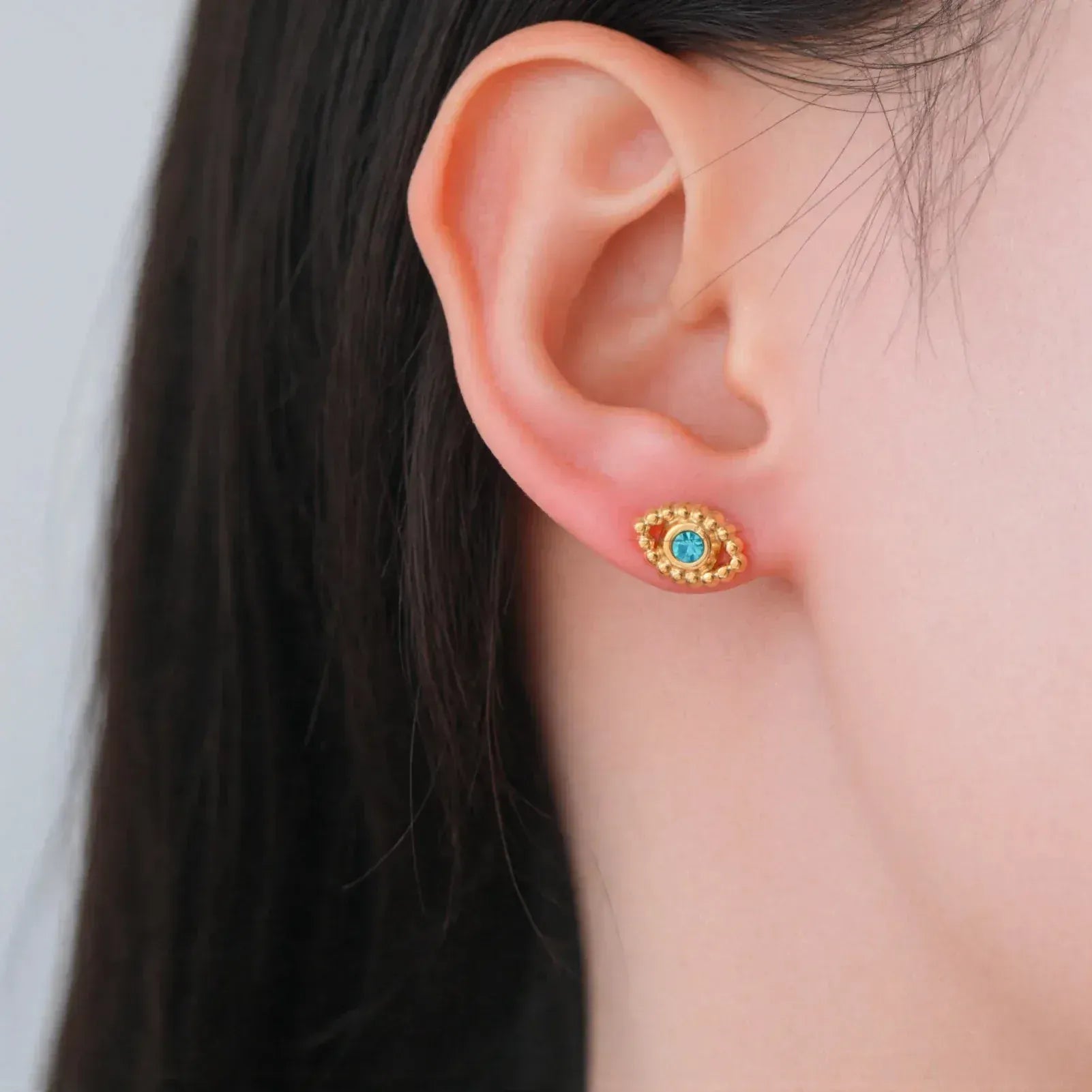 Close-up of an ear wearing a gold earring with a blue gemstone on a neutral background