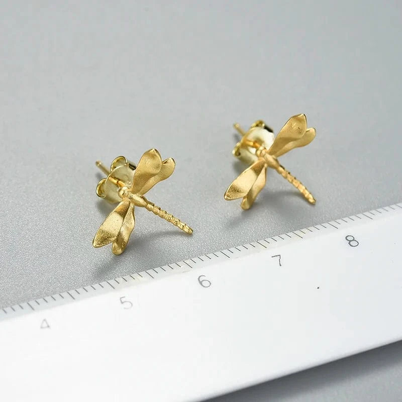 Gold dragonfly-shaped earrings on a gray surface with a ruler for scale.
