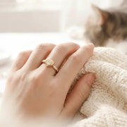 Hand wearing a silver ring with a paw print design, resting on a soft surface with a blurred cat in the background.