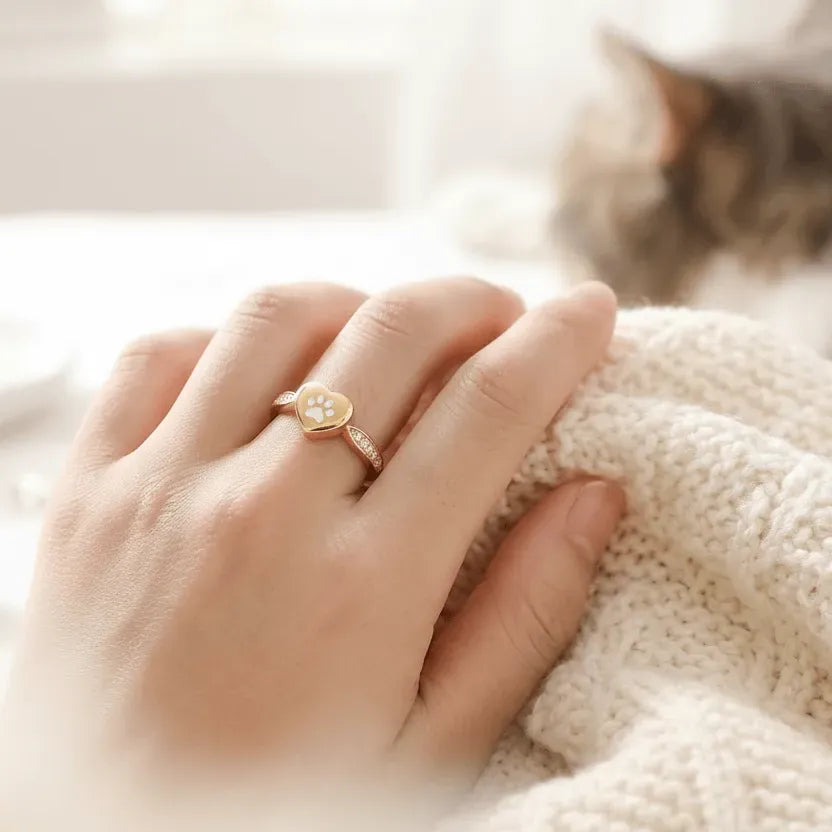 Hand wearing a silver ring with a paw print design, resting on a soft surface with a blurred cat in the background.