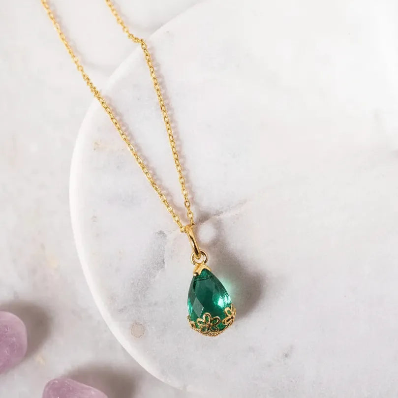 A gold chain necklace with a teardrop-shaped green gemstone pendant, styled on a white marble slab with purple tumbled stones nearby.