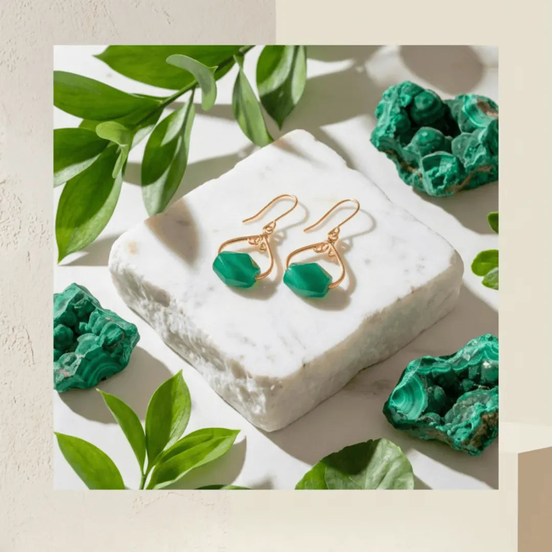 Gold earrings with green stones on a white stone surface surrounded by green leaves and crystals.