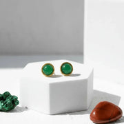 Gold earrings with green gemstones on a white stand, surrounded by natural elements.