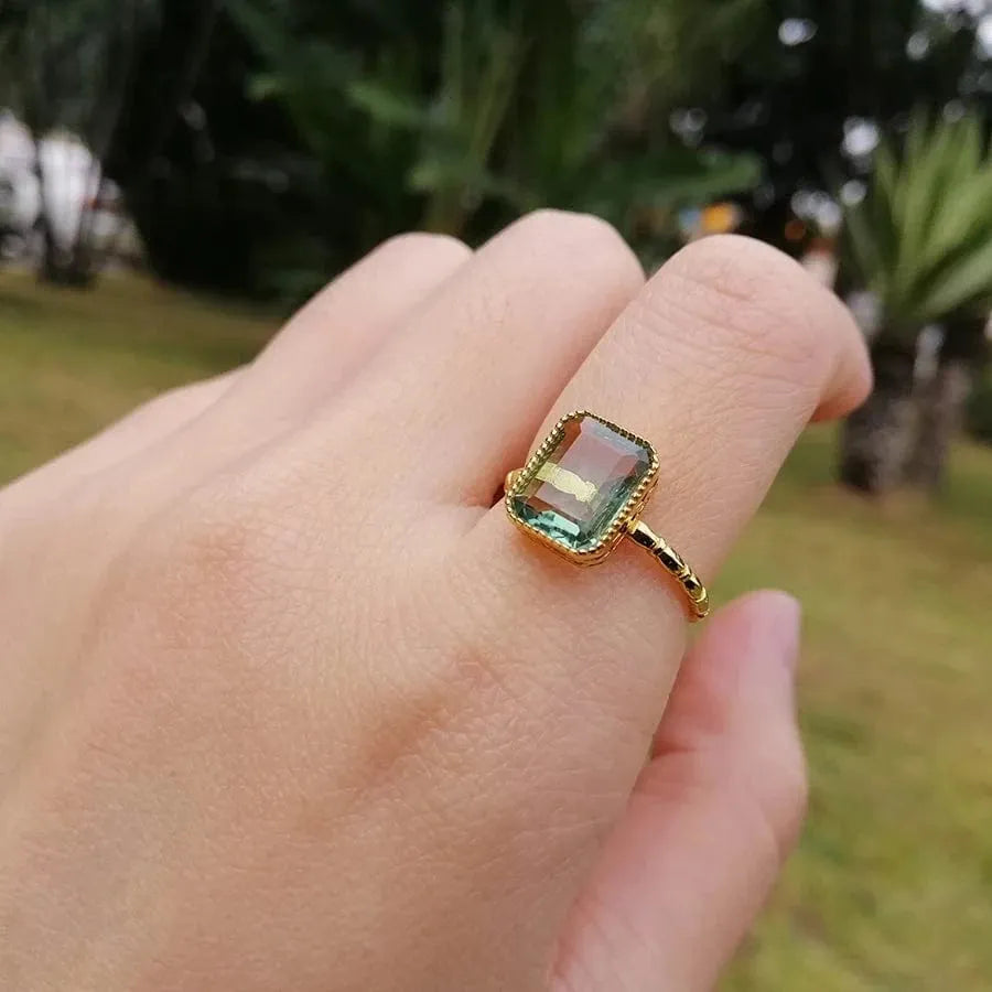 Gold ring with a green gemstone on a finger against a blurred outdoor background