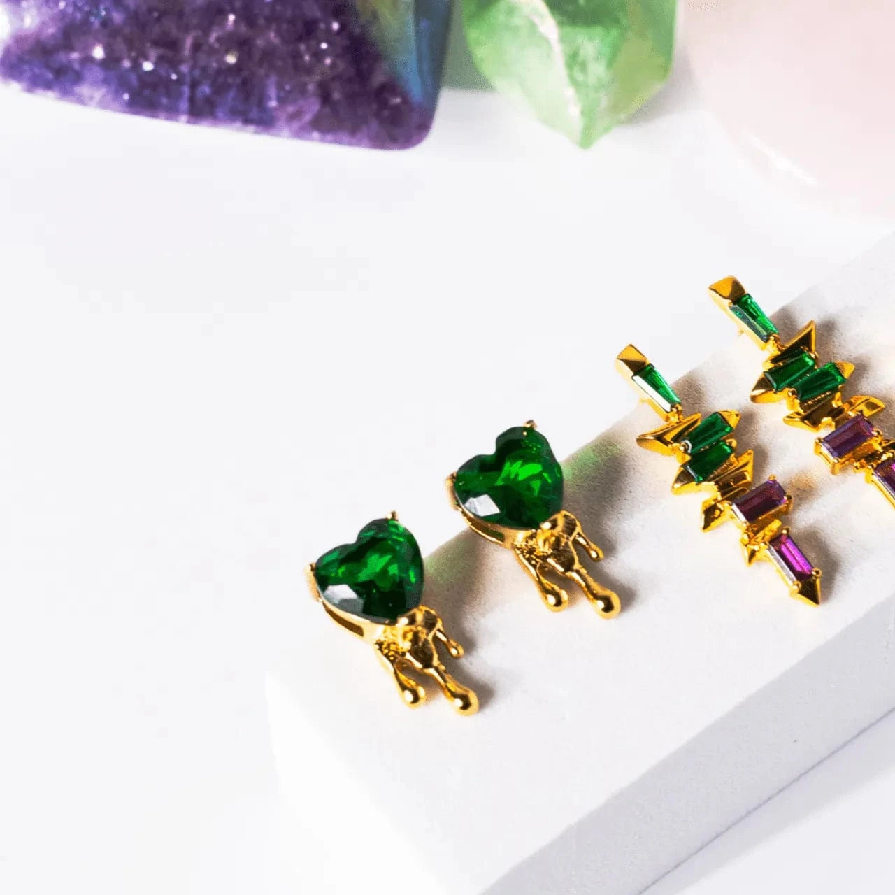 Gold and green gemstone earrings on a white surface with purple and green crystals in the background.