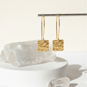 Gold square earrings on a white stand with a light background